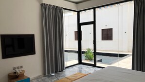 Habitación de hotel con cama, televisor montado en la pared, mesita de noche pequeña con teléfono y puerta corrediza de vidrio que da a un área exterior con piscina.