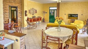 A dining area with a round table, chairs, and a vase of flowers. There is a sofa and a television in the background.