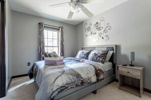 Une chambre à coucher comprenant un lit, une table de chevet, un ventilateur de plafond, une fenêtre avec des rideaux et un mur décoré de motifs floraux.