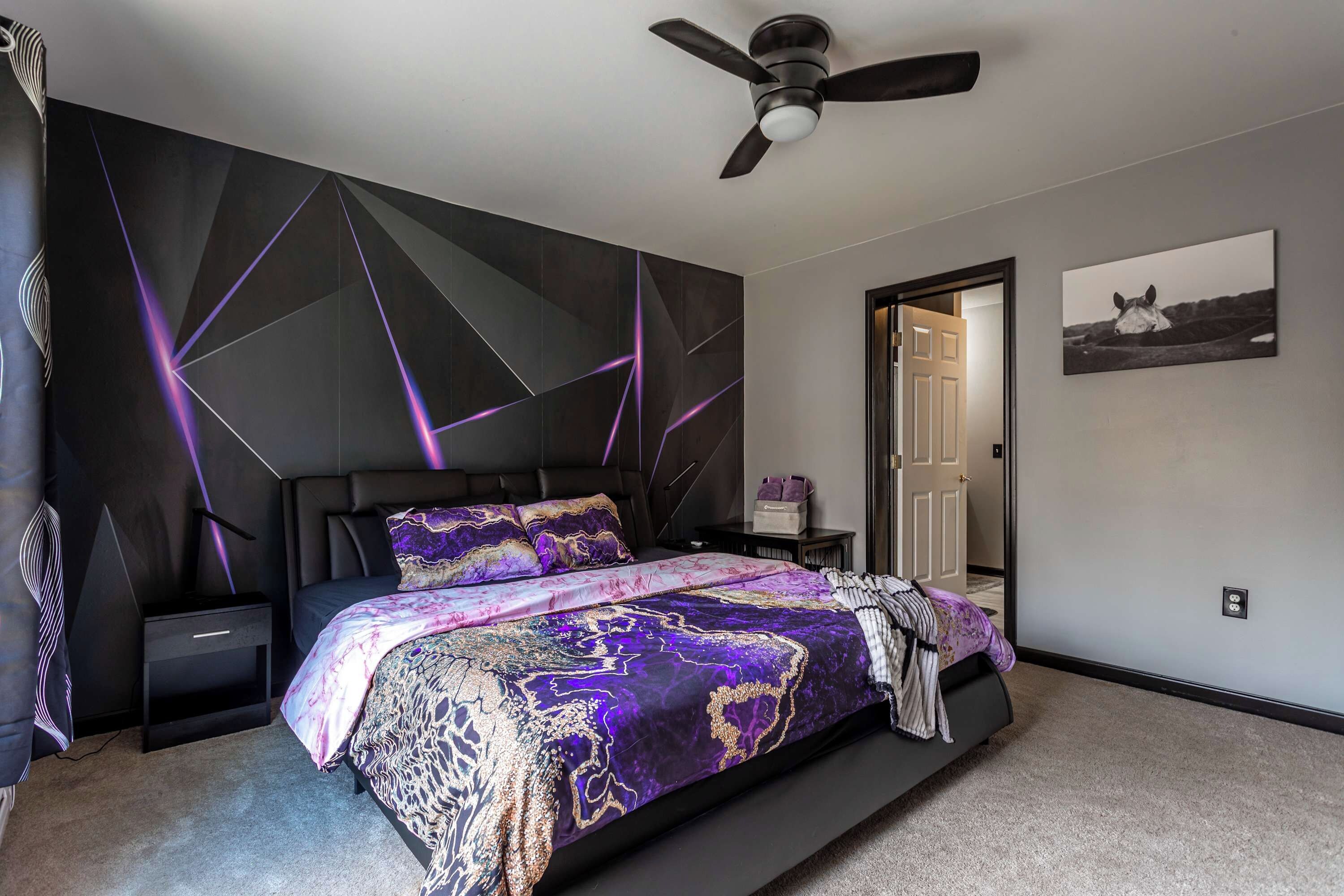 Een slaapkamer met een modern design, met een bed met een beddengoed met een patroon, een plafondventilator en een deur die naar een andere kamer leidt.