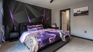 A bedroom with a modern design, featuring a bed with a patterned bedspread, a ceiling fan, and a door leading to another room.