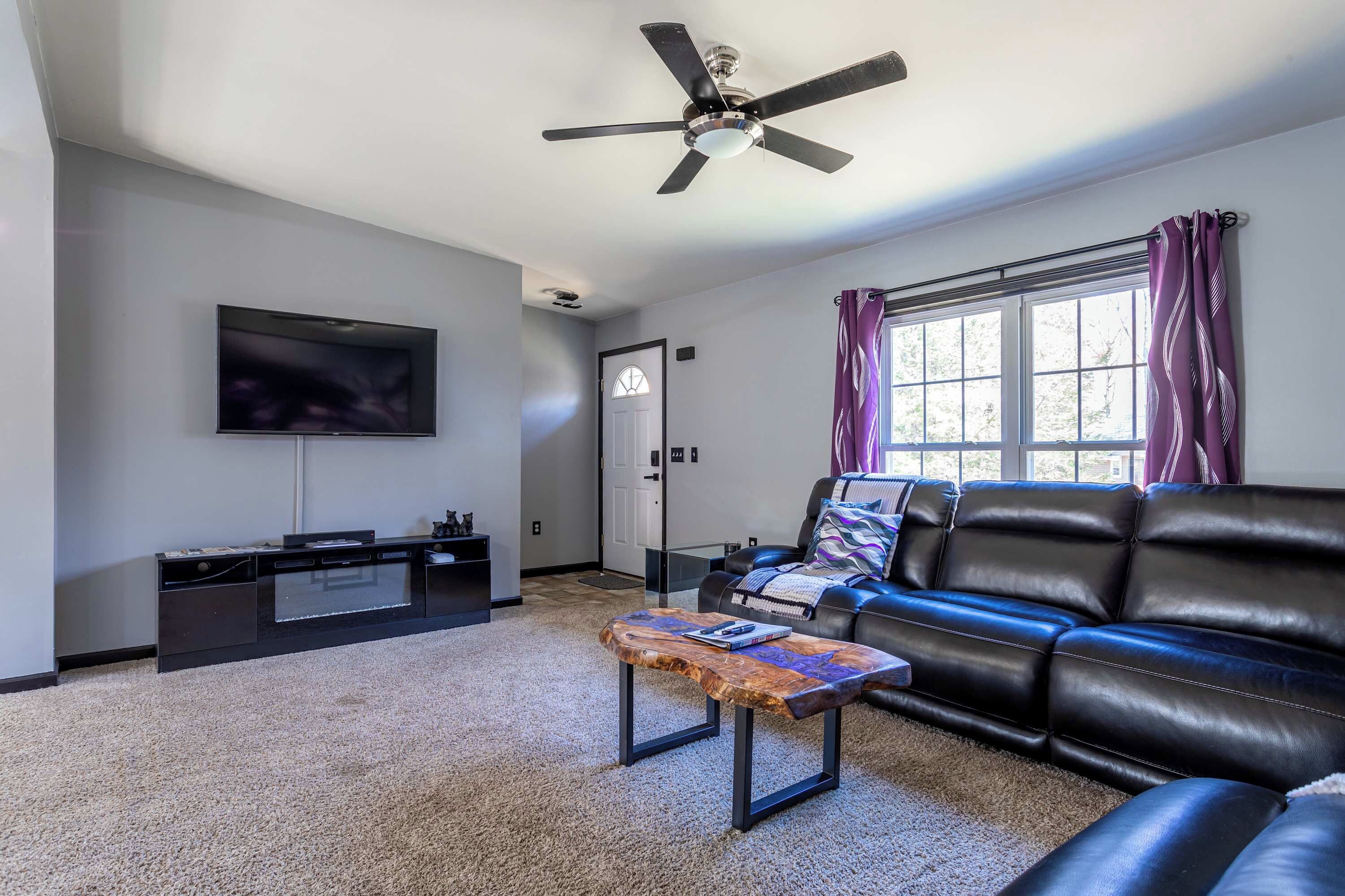 A living room with a flat-screen TV, a ceiling fan, a black leather sofa, a coffee table, and a window with purple curtains.