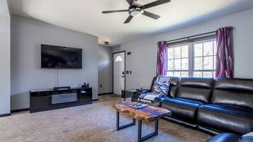 A living room with a flat-screen TV, a ceiling fan, a black leather sofa, a coffee table, and a window with purple curtains.
