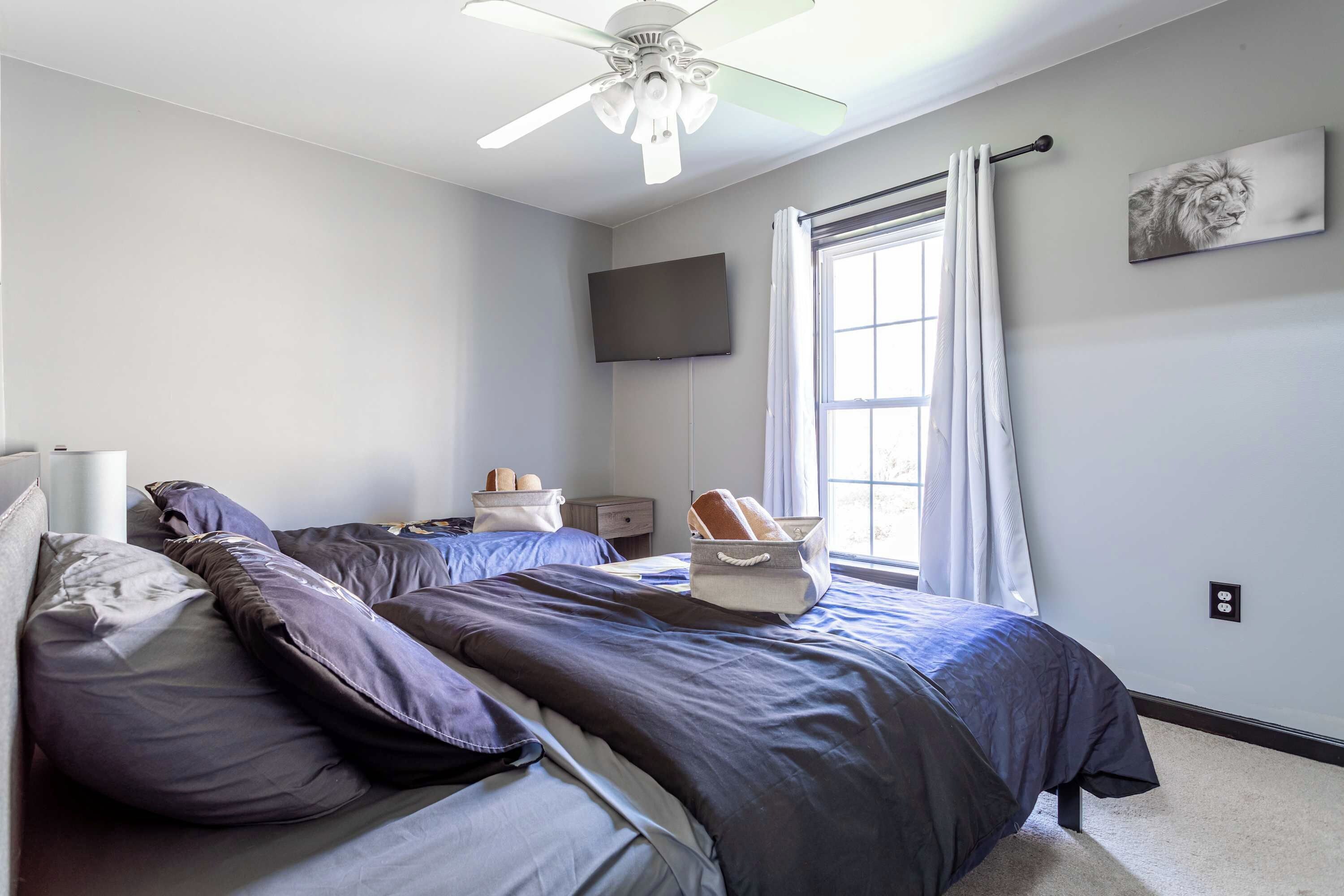 A bedroom with a bed, pillows, a nightstand, a flat-screen TV, a window with curtains, and a ceiling fan.