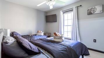 A bedroom with a bed, pillows, a nightstand, a flat-screen TV, a window with curtains, and a ceiling fan.