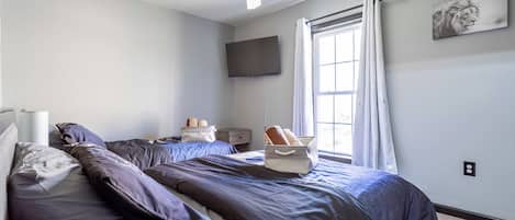 A bedroom with a bed, pillows, a nightstand, a flat-screen TV, a window with curtains, and a ceiling fan.