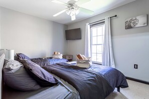 Un dormitorio con cama, almohadas, mesita de noche, televisor de pantalla plana, ventana con cortinas y ventilador de techo.