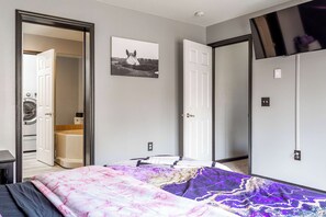 Un dormitorio con una cama, una puerta, un televisor montado en la pared y un cuadro enmarcado.