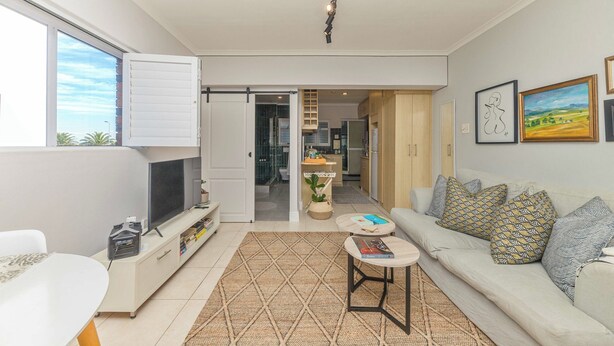 A modern living room with a sofa, coffee table, TV, and a kitchen in the background. - Mouille Point Studio including welcome basket (Cape Town)