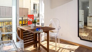 A room with a wooden table, a clear plastic chair, a TV mounted on the wall, and a window overlooking a balcony.