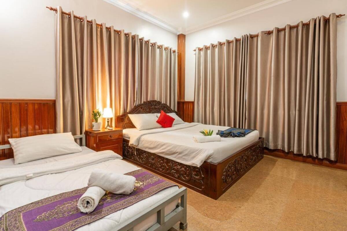Quarto de hotel com duas camas, cabeceira de madeira, mesa de cabeceira com abajur e cortinas bege.