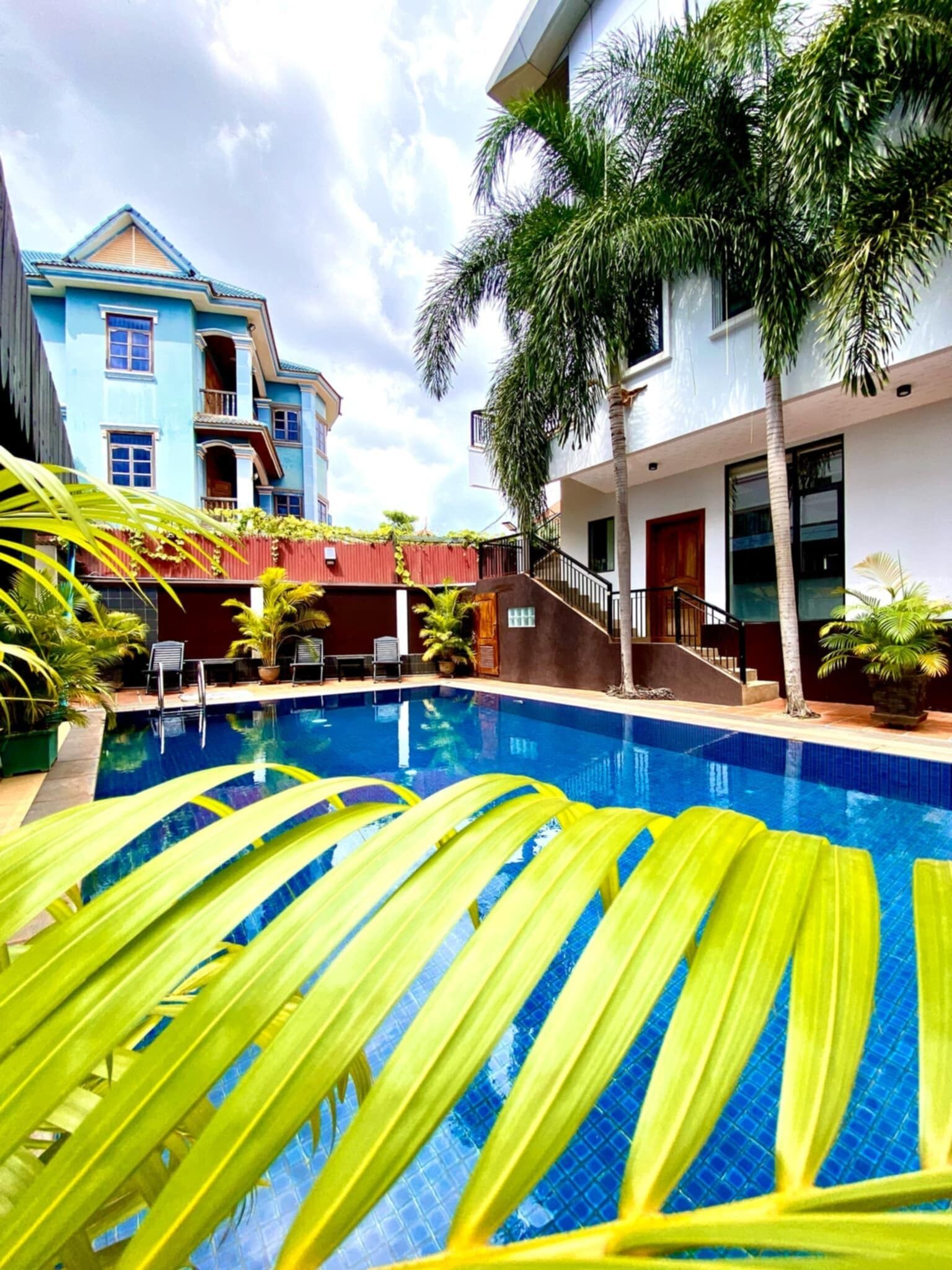 A swimming pool with a clear blue water surface, surrounded by palm trees and a building with balconies.