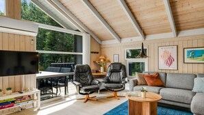 Un salon spacieux avec un plafond haut, des murs en bois, une grande fenêtre, un téléviseur à écran plat, un canapé gris, une table basse et une bibliothèque.