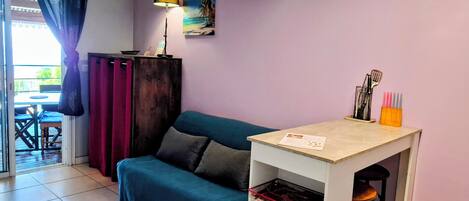 A room with a teal sofa, a small table with a lamp, a painting on the wall, and a view of the outdoors through a window.
