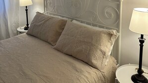Un lit bien fait, recouvert d’une couvre-lit à motifs et agrémenté de deux oreillers, est flanqué de deux tables de chevet sur lesquelles reposent des lampes. Un tableau représentant un arbre, encadré, est accroché au mur au-dessus du lit.