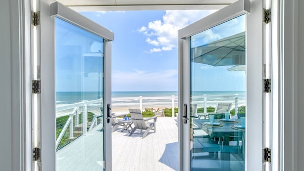Puerta de vidrio que da a una terraza con asientos exteriores y vista a la playa.