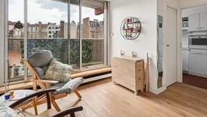 Un appartement moderne doté d’une grande porte-fenêtre coulissante donnant sur un balcon, d’une petite cuisine équipée et d’un coin salon avec une chaise et une table.