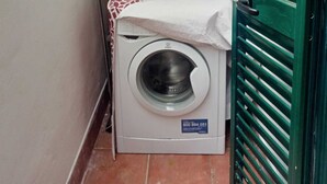 Een kleine wasruimte met een wasmachine, een fles wasmiddel en een blauwe bak op een tafel.