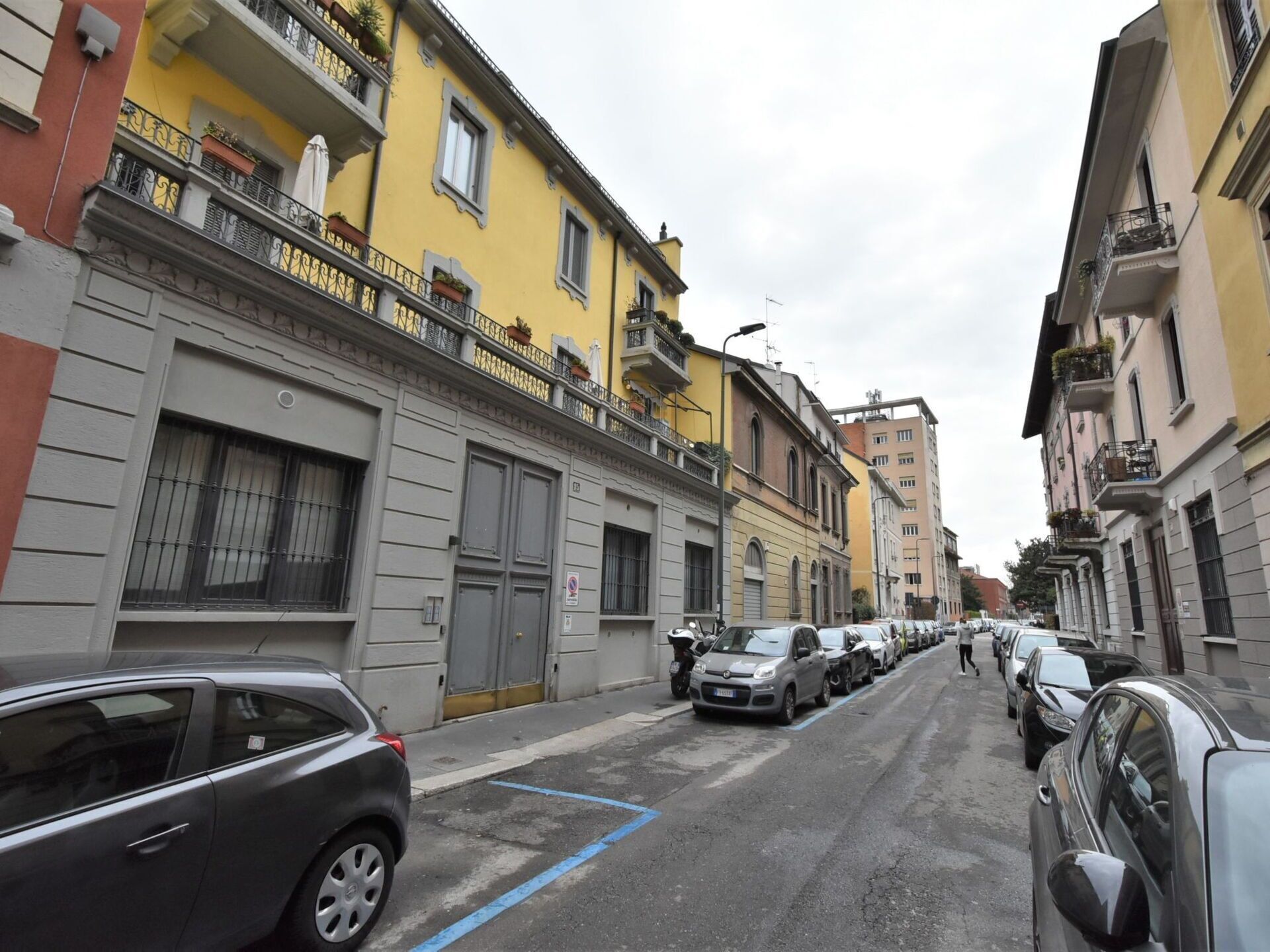 Une rue étroite bordée d’immeubles à plusieurs étages, de voitures garées et d’un piéton.
