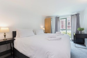 A hotel room with a bed, bedside table, lamp, and a view of buildings through the window. - Raleigh Square-2BR Ground-Floor Apt in Nottingham (Nottingham)