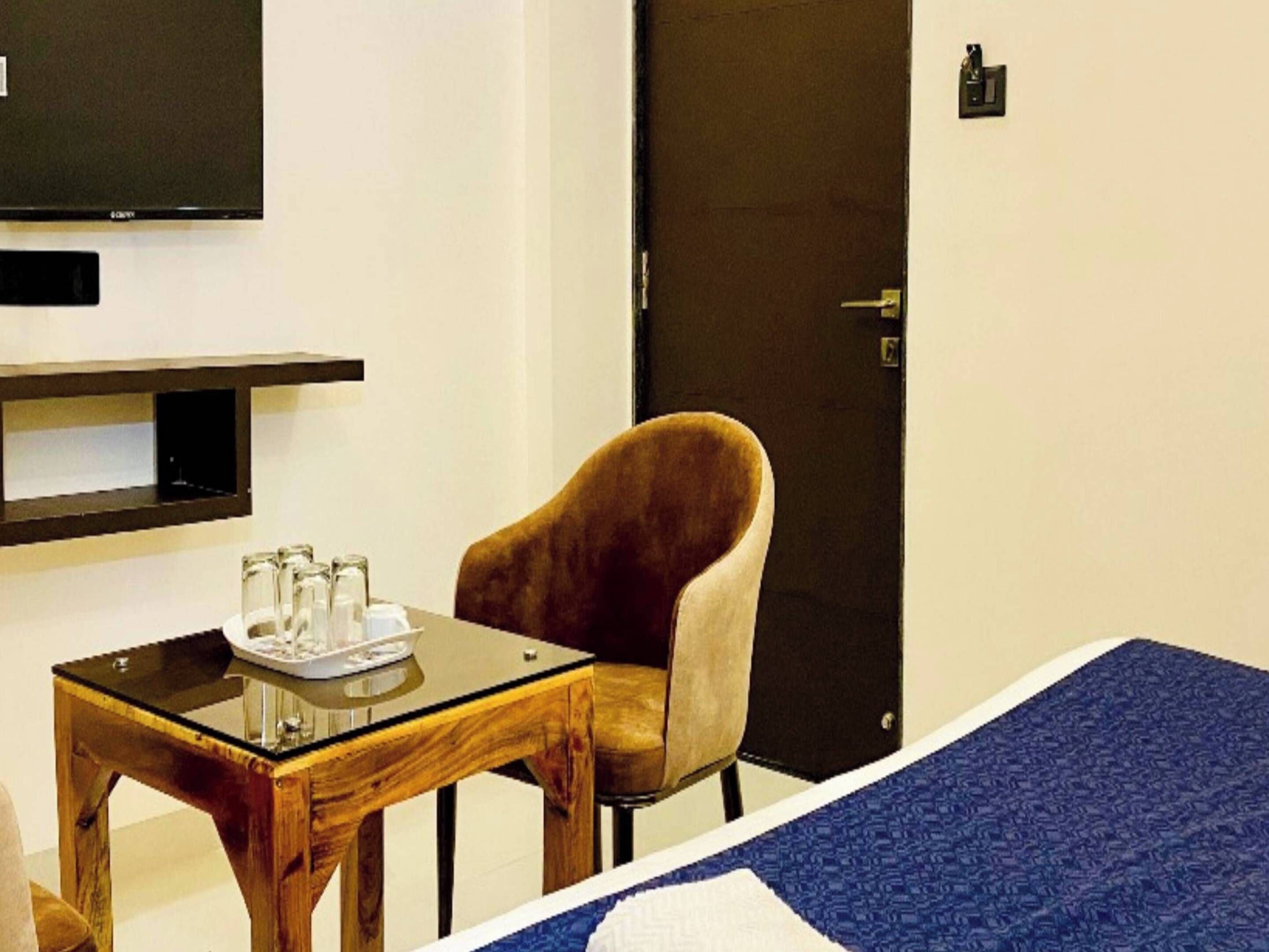A hotel room with a bed, a wooden side table with glasses and a tray, a brown armchair, and a television mounted on the wall.
