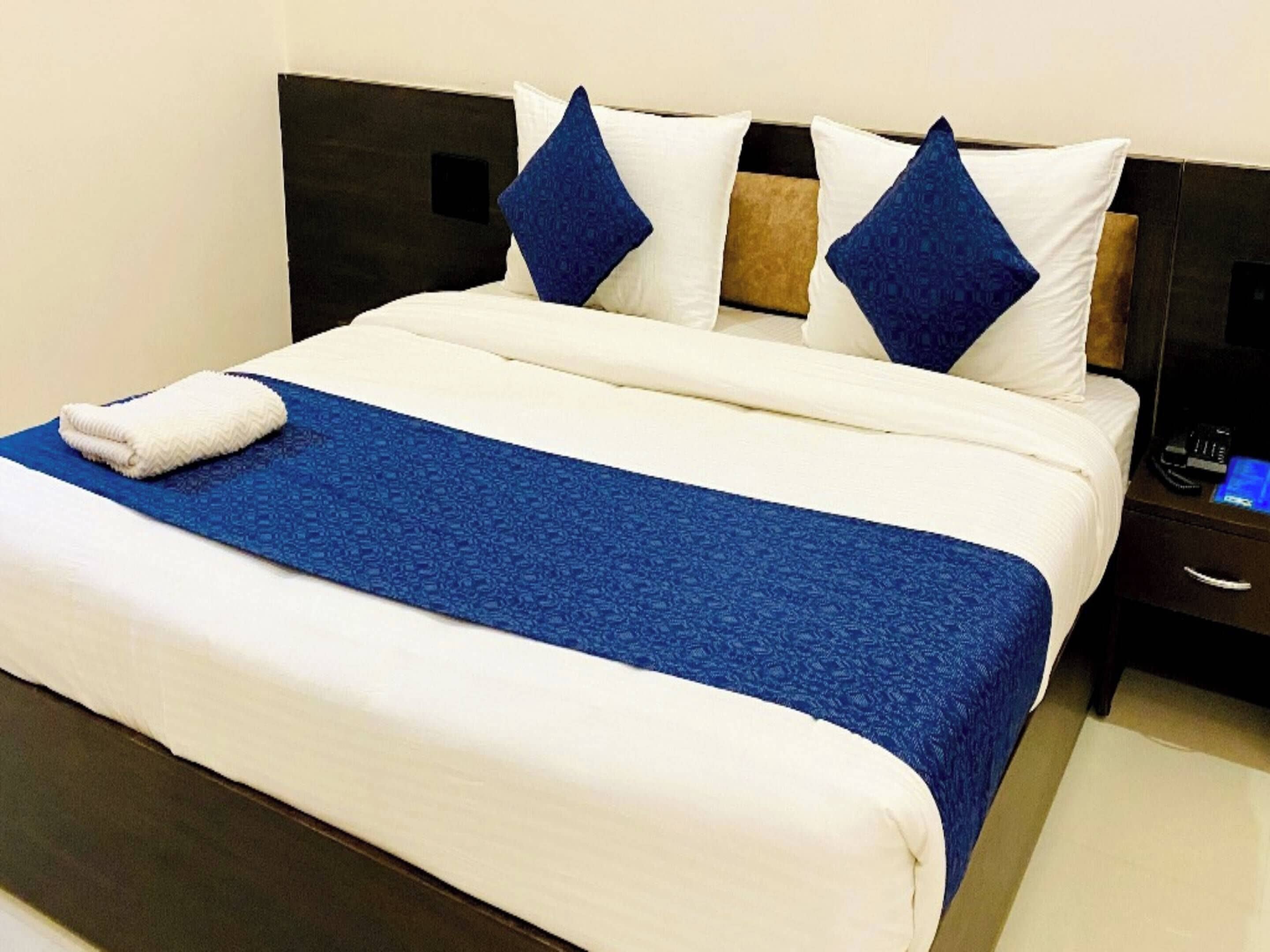 A neatly made bed with a blue bedspread and white pillows, a white towel on the bed, and a nightstand with a phone and a remote control.