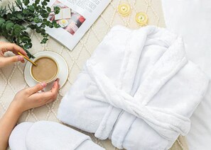 Henkilö, jolla on kädessään teekuppi, päällään valkoinen kylpytakki ja hänellä on sylissään lehti, ja hän seisoo tai istuu pinnalla, jossa on selkeä kuvio tai rakenne.