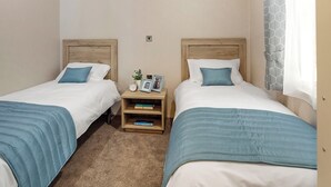 Ein Doppelzimmer mit zwei Betten, jeweils mit Holz Kopfteilen, weißer Bettwäsche und blauen Ziertelementen. Ein Nachttisch beherbergt eine Lampe, eine kleine Pflanze und gerahmte Fotos. Die Wände sind beige und ein Fenster ist mit einem gemusterten Vorhang versehen.