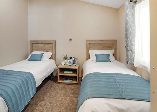 Ein Doppelzimmer mit zwei Betten, jeweils mit Holz Kopfteilen, weißer Bettwäsche und blauen Ziertelementen. Ein Nachttisch beherbergt eine Lampe, eine kleine Pflanze und gerahmte Fotos. Die Wände sind beige und ein Fenster ist mit einem gemusterten Vorhang versehen.