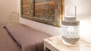 A bed with a textured blanket, a rustic wall with a distressed painting, and a decorative lantern on a white table.