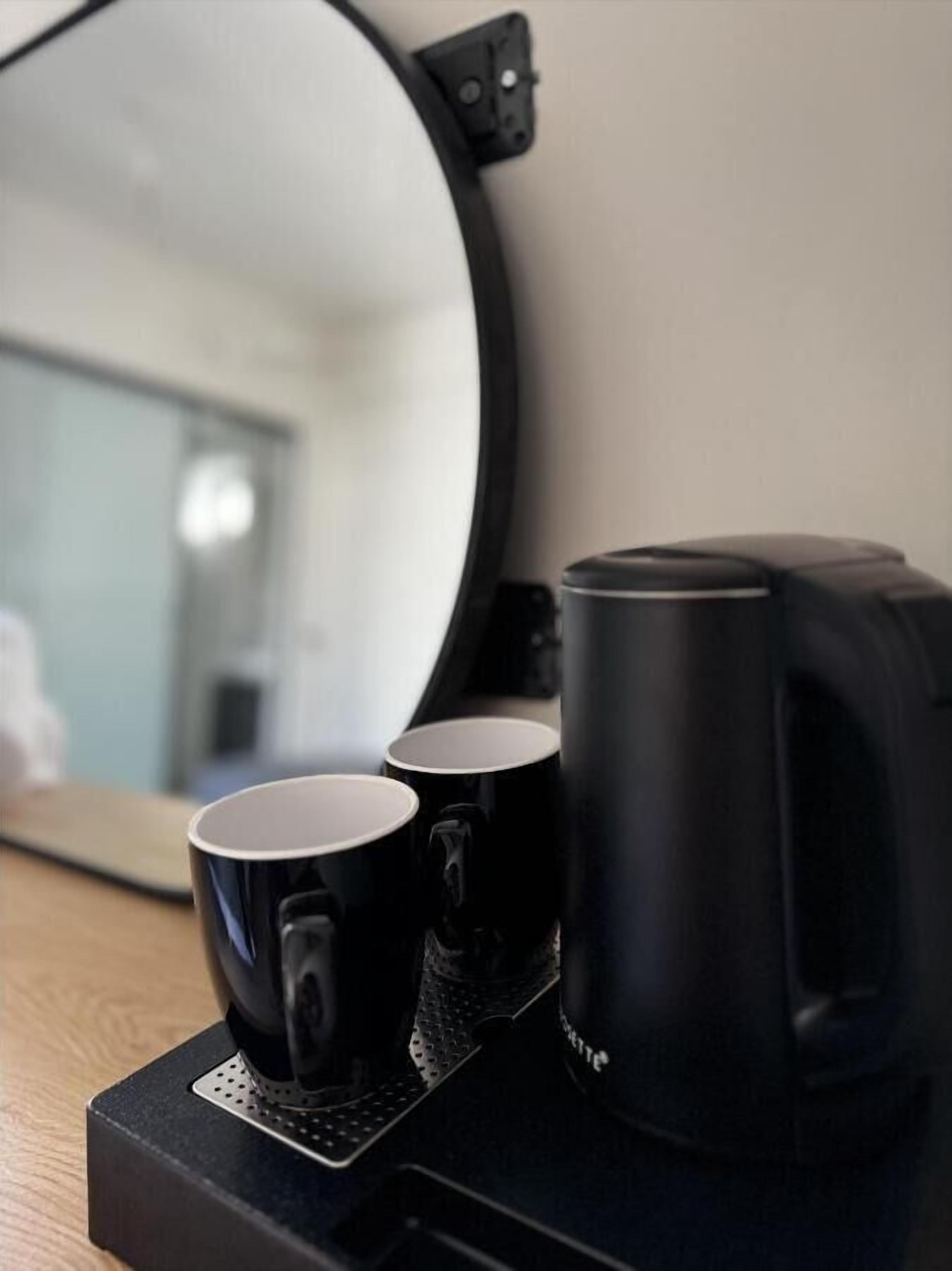 Una cafetera con dos tazas negras sobre un posavasos, un espejo que refleja una habitación, y un aplique de pared.