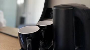 A coffee maker with two black mugs on a coaster, a mirror reflecting a room, and a wall-mounted light fixture.