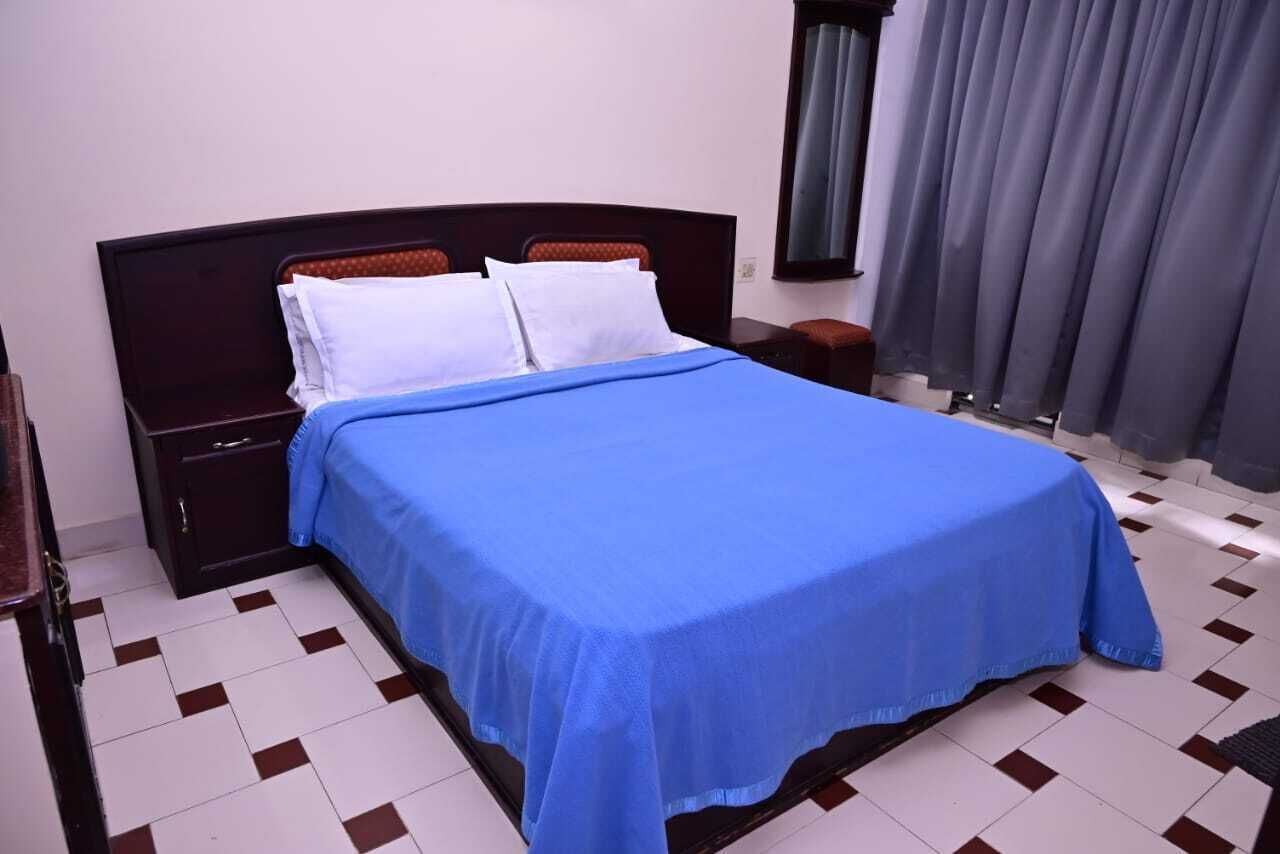 A neatly made bed with a blue bedspread and white pillows, a dark wooden headboard, a nightstand with a drawer, and a window with grey curtains.