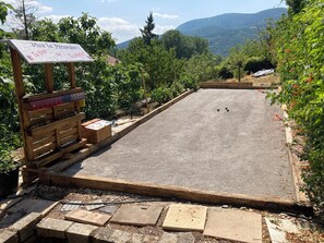 A pétanque court with a wooden bench and a sign, surrounded by greenery and a mountain view.
