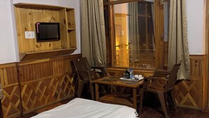 A hotel room with a bed, a wooden TV stand, a table with chairs, and a large window with curtains. - Woodward Magique kasol (Bhuntar)