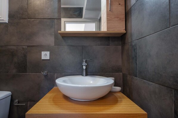 A modern bathroom with a large white vessel sink on a wooden countertop, dark stone walls, and a wooden medicine cabinet above the sink. - Villa Spirida at Drakopoulata (Kefalonia)