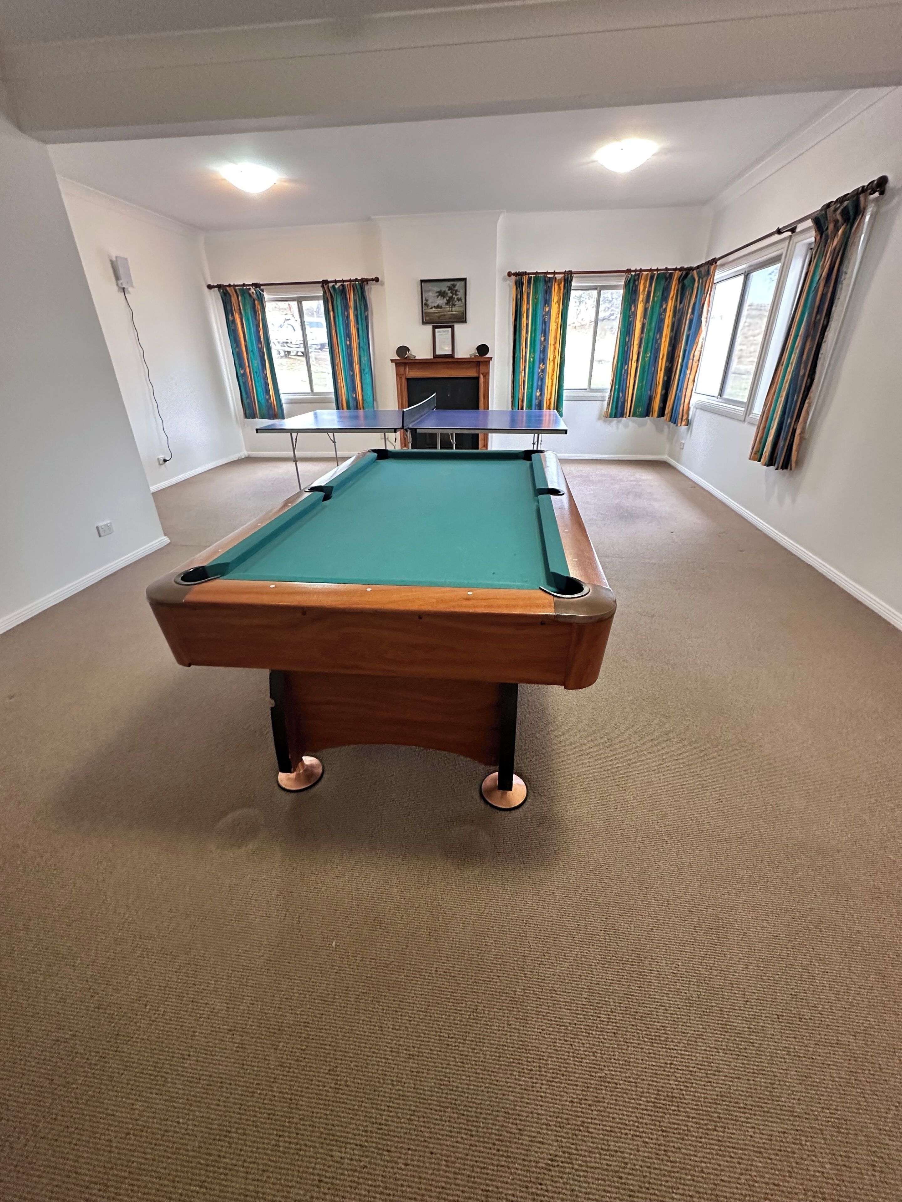 Une table de billard dans une pièce dont le sol est recouvert de moquette, les murs sont blancs et de grandes fenêtres sont ornées de rideaux.