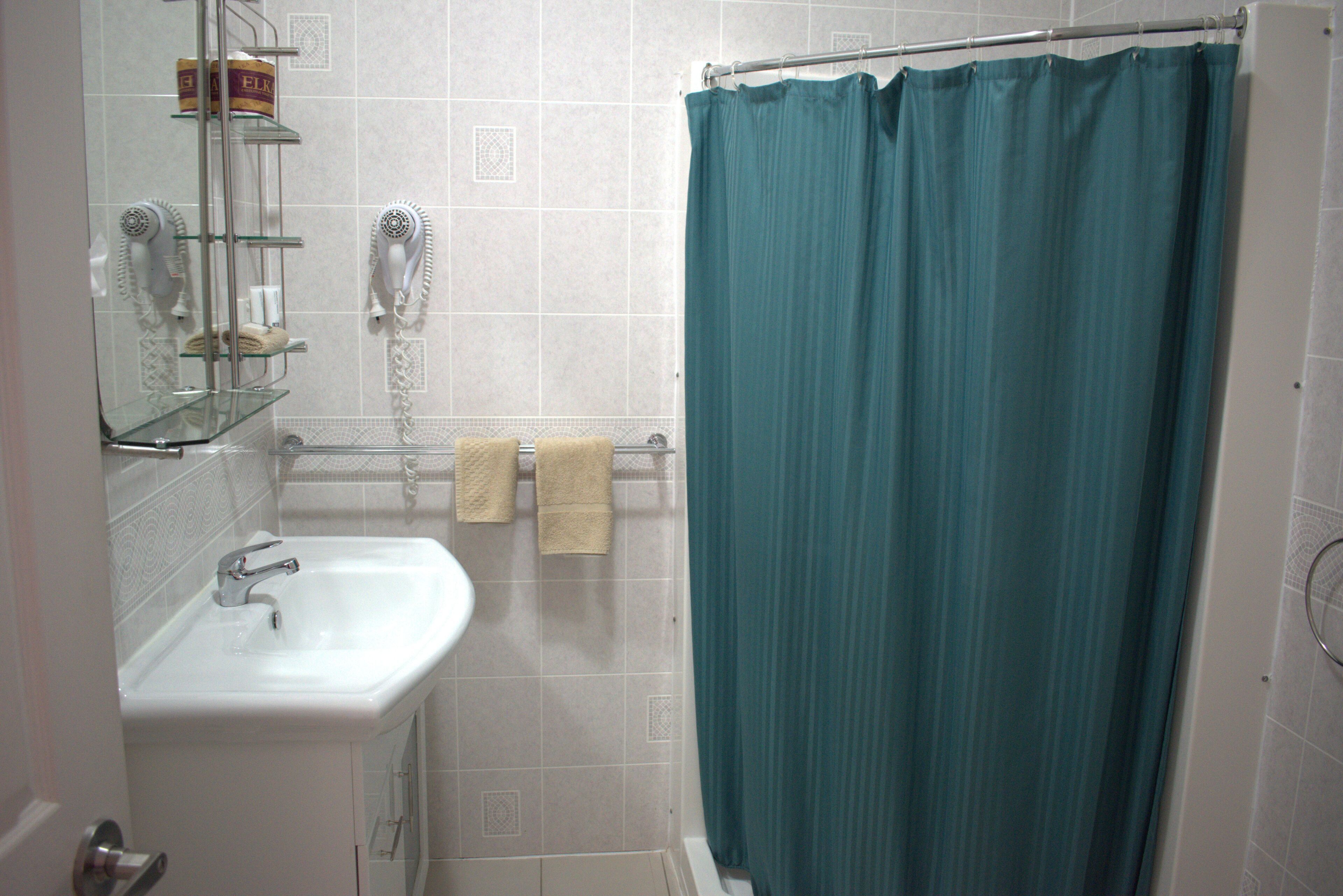 Une salle de bain avec un lavabo blanc, une étagère en verre où sont rangés les articles de toilette, un porte-serviettes avec deux serviettes et un rideau de douche de couleur turquoise.