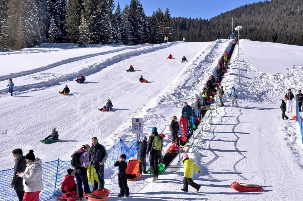 A snowy ski slope with people sledding and standing by the side.