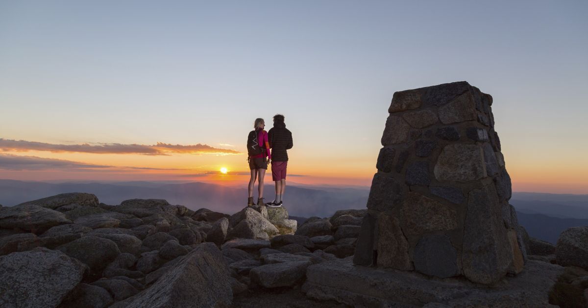 Due persone su uno sperone roccioso, con un monumento in pietra in primo piano e un tramonto sullo sfondo.