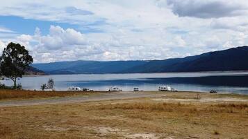 Vista de un lago con vehículos recreativos estacionados a lo largo de la orilla, una persona de pie en primer plano y montañas al fondo.