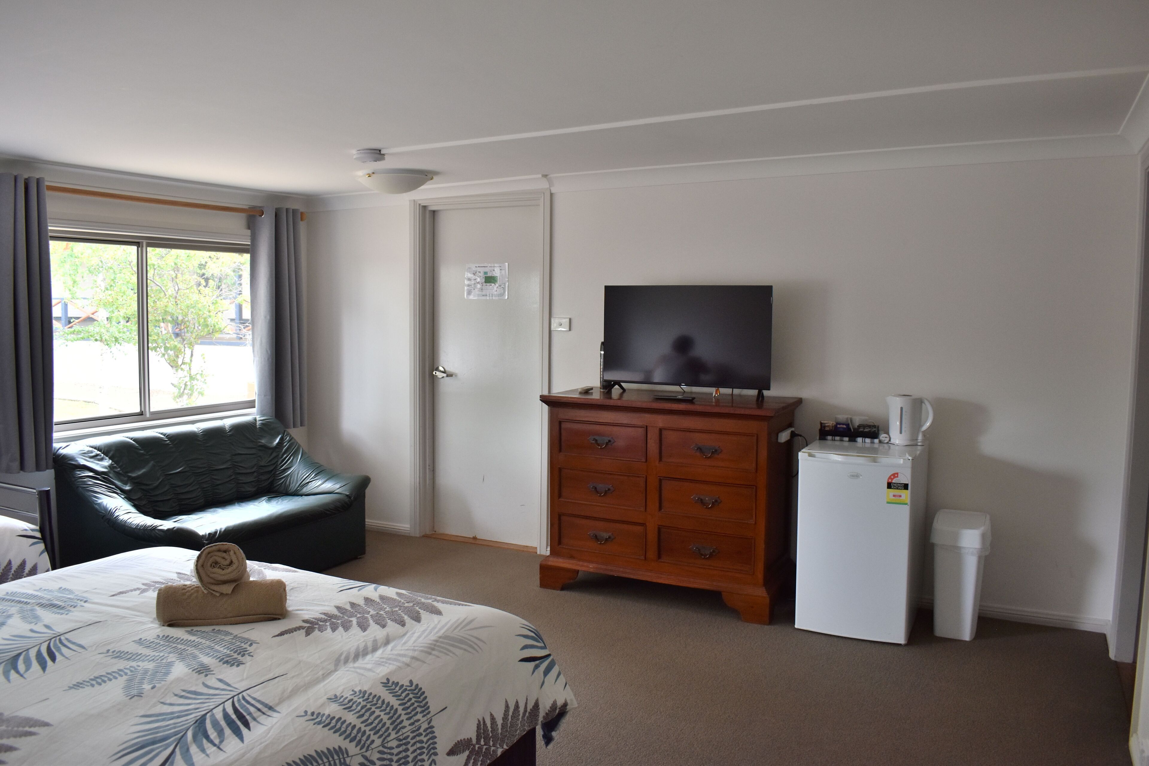 A hotel room with a bed, a television, a wooden dresser, a refrigerator, and a window with curtains.