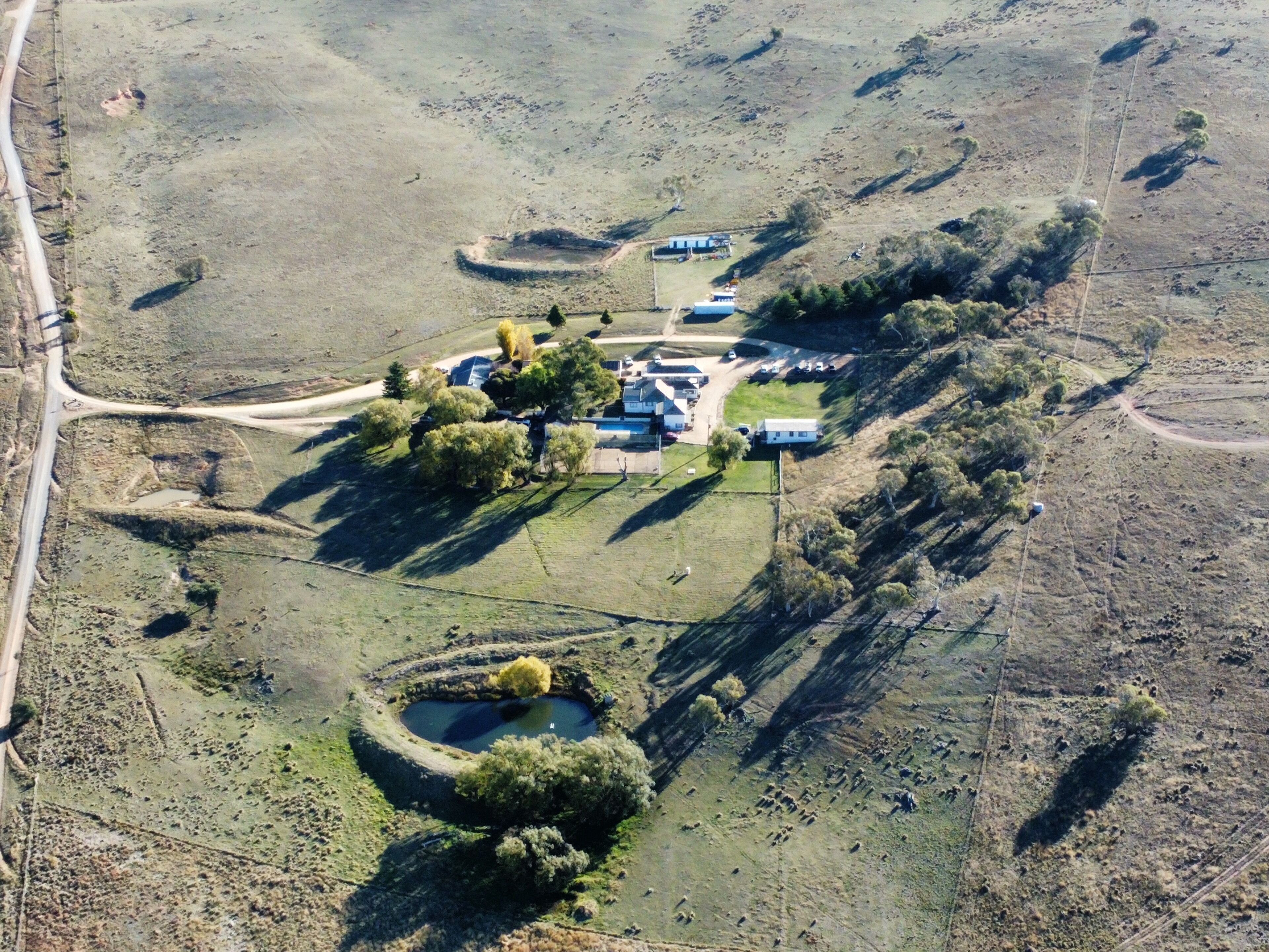 An aerial view of a rural property with a central building surrounded by trees, a small pond, and a road to the left.