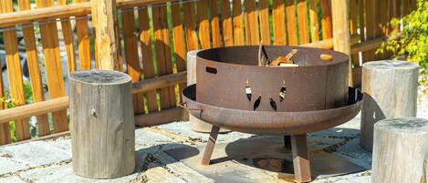Eine Metall-Feuerstelle mit rustikalem Design, platziert auf einer Steinterrasse mit Holzstützen.