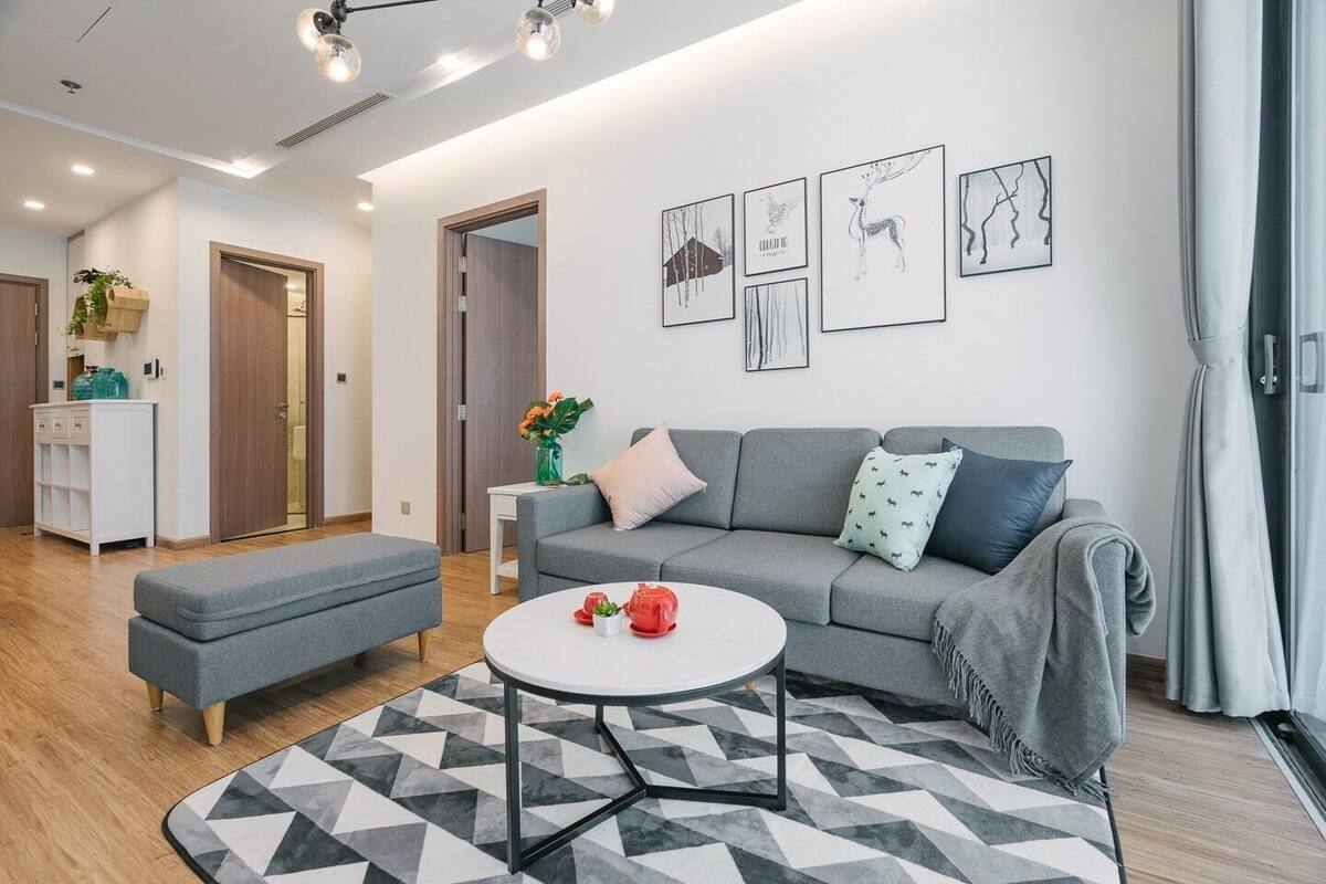 A modern living room with a grey sofa, a round coffee table, and a geometric rug. There are framed artworks on the wall and a plant on the side table.