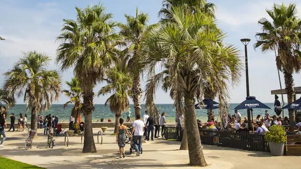 A beachside promenade with palm trees, people walking, and beach umbrellas. - Studio Apartment in St Kilda next to Luna Park (St Kilda)