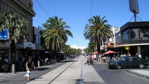 A street with palm trees, shops, and a tram. - Studio Apartment in St Kilda next to Luna Park (St Kilda)