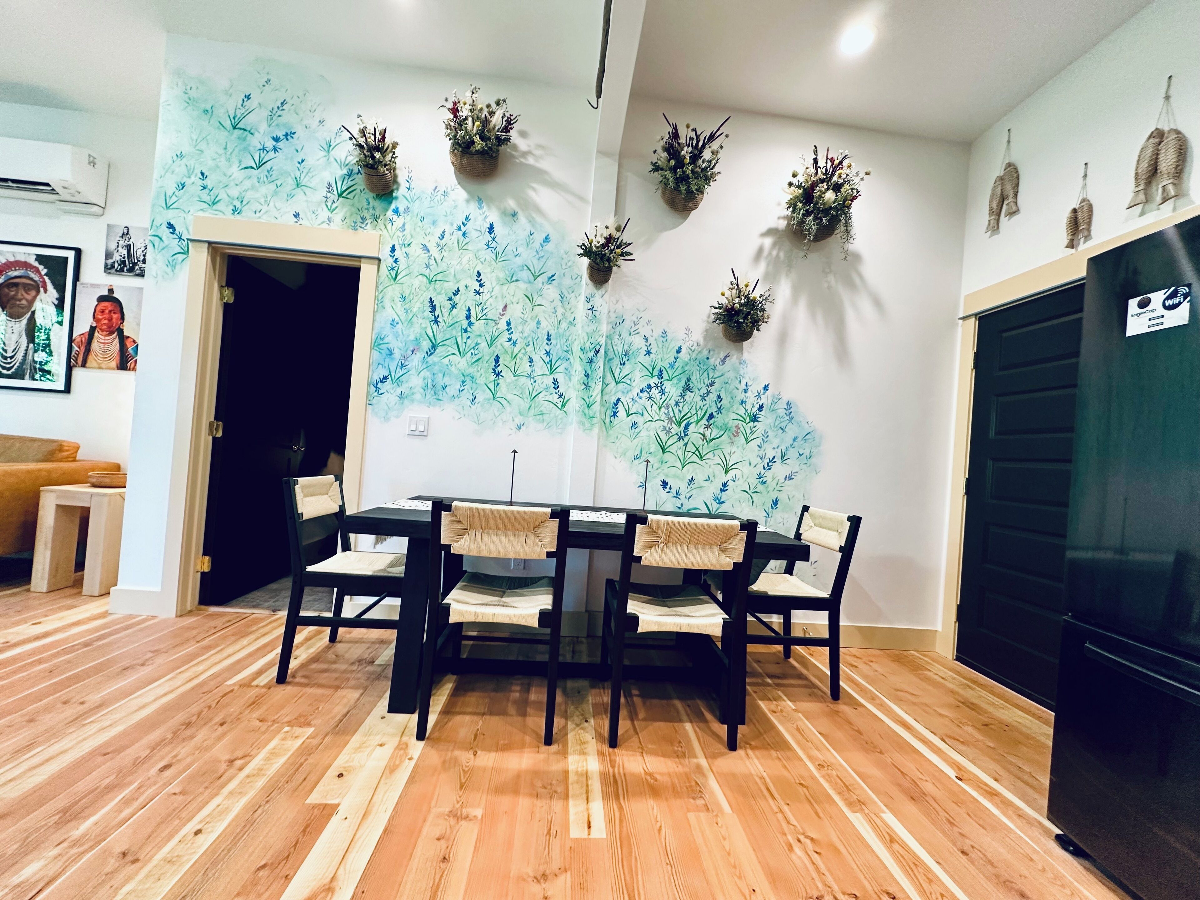 A dining area with a wooden floor, a black table, and four chairs with woven seat covers. There are wall-mounted planters with greenery.