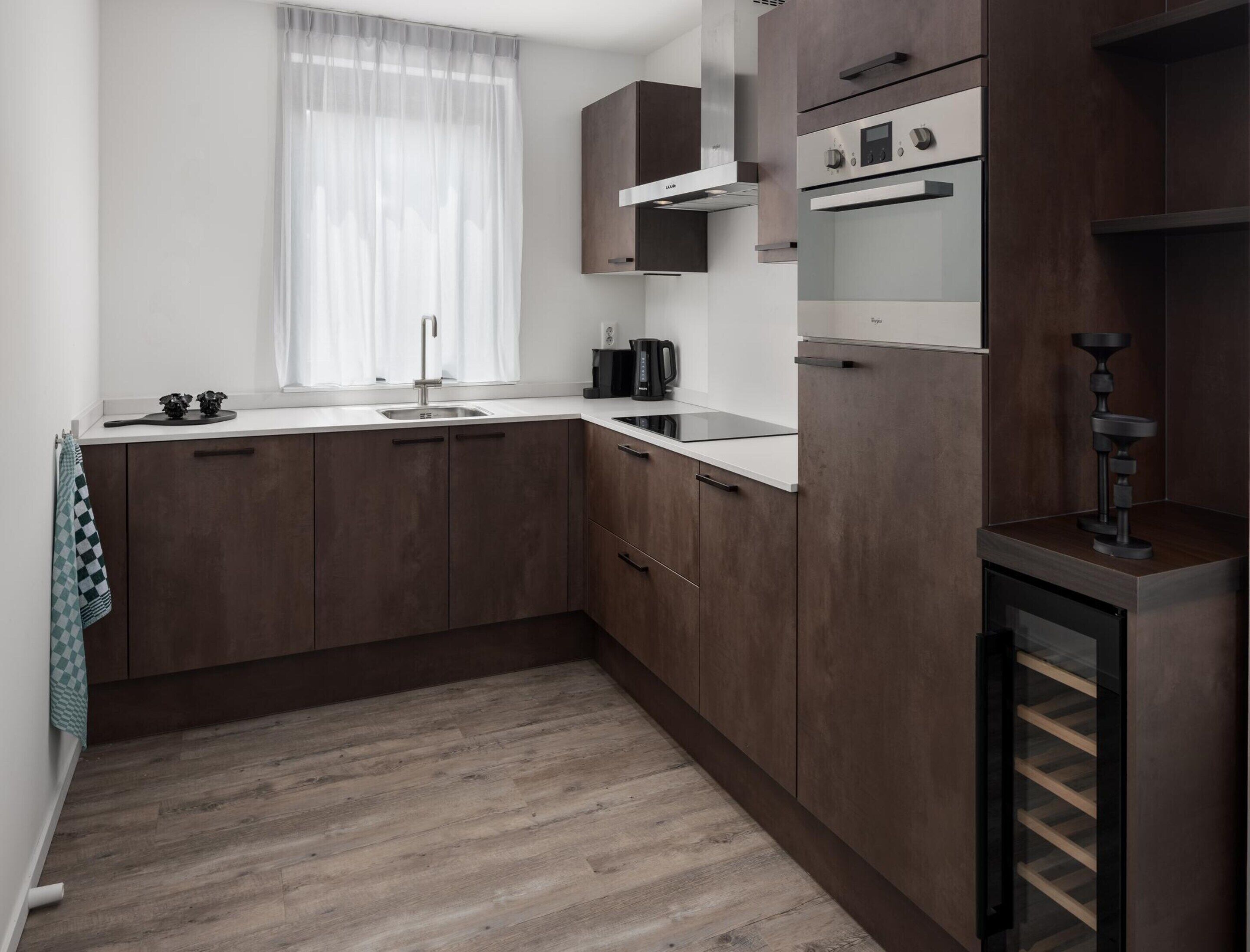 A compact kitchen with dark wooden cabinets, a white countertop, a stainless steel sink, and a built-in oven.