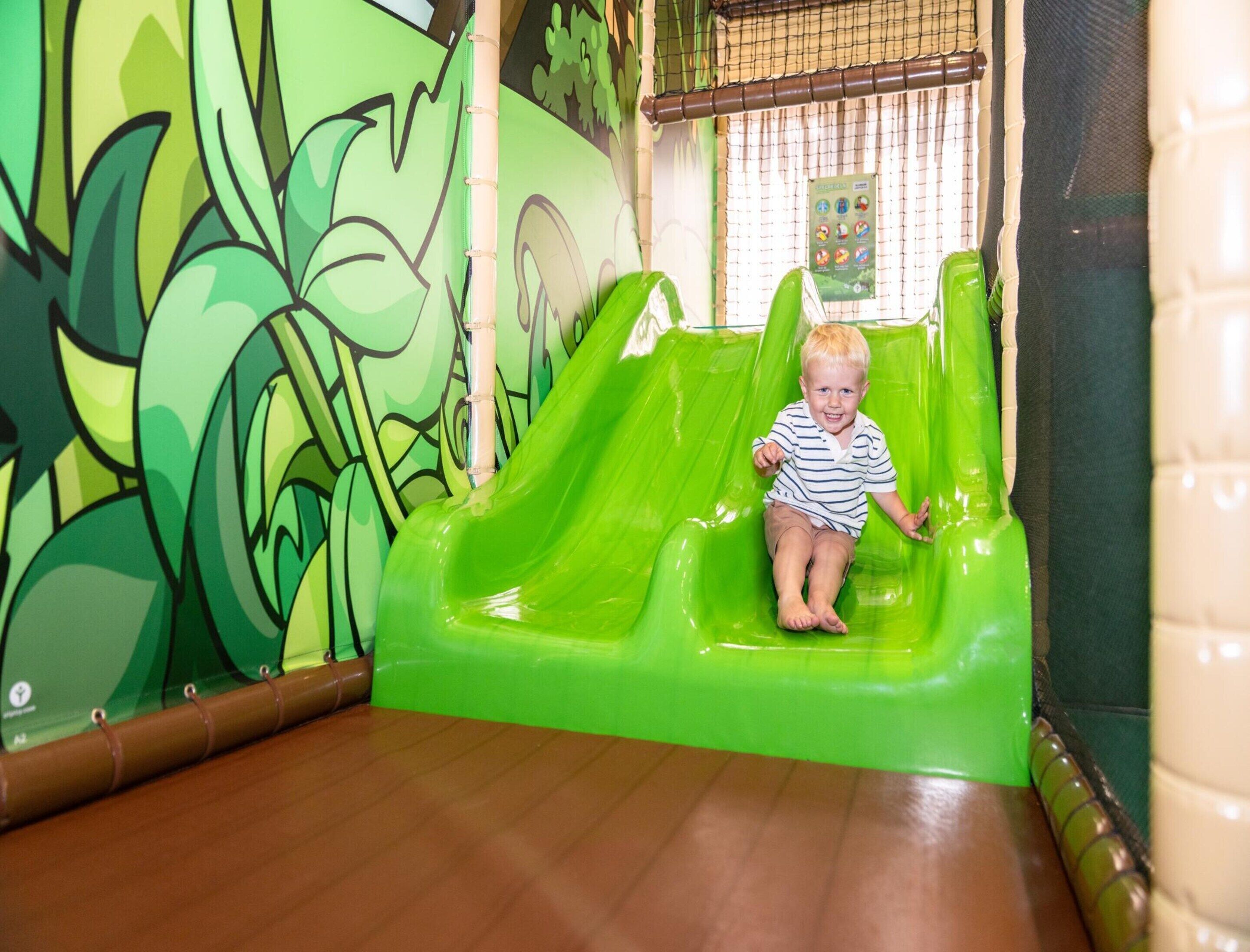 A child on a green slide in an indoor play area with a mural of green foliage on the wall.